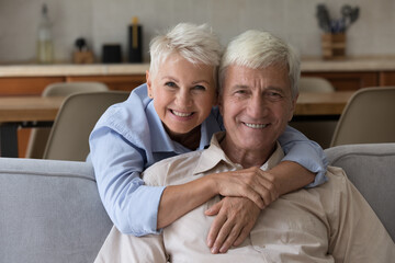 Aged couple smile pose for camera, loving wife piggyback husband seated on sofa at home looking at...