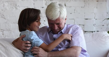 Happy active granddad and cute energetic grandson kid tickling each other on couch, playing at home, enjoying active games, leisure time together, laughing, giggling, having fun