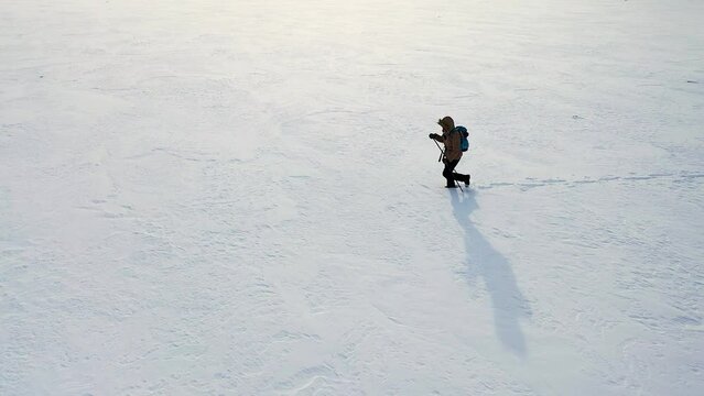 Lonely hiker in down jacket with backpack and ski poles walk through snow - covered wasteland, drone aerial side view.