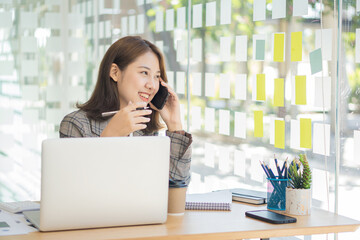 Obraz premium Portrait of Asian businesswoman sitting at a desk and working on a laptop computer with a phone call and financial graph documents on the table.