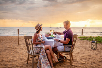 Couple in love drinking champagne wine on romantic dinner at sunset on the beach with yachts on background. Young couple celebration anniversary, clink glasses at served table on sea sandy coastline.