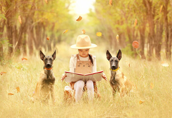 A little girl sits in the forest and reads a book, she is guarded by malinois puppies, autumn nature