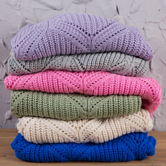 six colored knitted sweaters, lie one on top of the other, against the background of a gray wall