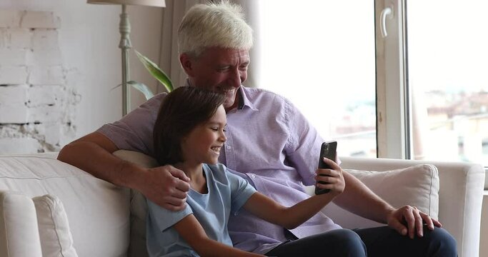 Cheerful gen Z grandson boy and happy grandpa recording self video on smartphone for social media, holding mobile phone, looking at screen, laughing, sitting on home sofa together