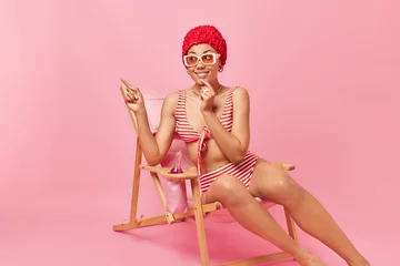 Tuinposter Pretty young woman dressed in striped swimsuit sunglasses and hat has cheerful dreamy expression points away into distance sits on deck chair shows something far away isolated over pink wall © Wayhome Studio