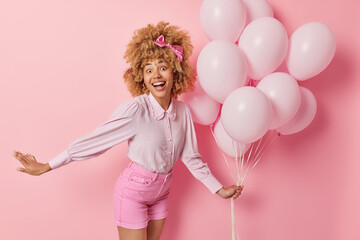 Positive surpised woman spends free time on welcome party holds bunch of inflated balloons wears blouse and shorts feels very happy dances against pink background. Birthday celebration concept