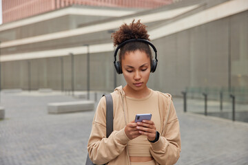 Outdoor shot of curly haired dressed in beige sportswear carries fitness mat listens music via...