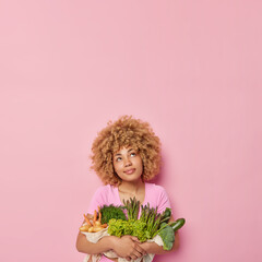 Vertical shot of thoughtful woman carries fresh vegetables eats healthy food full of vitamins wears casual t shirt isolated over pink background empty space for your advertisement. Gardening concept