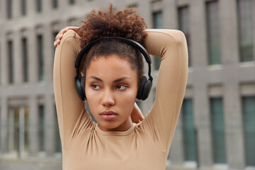 Athletic female model with curly hair stretches arms and looks thoughtfully into distance listens...