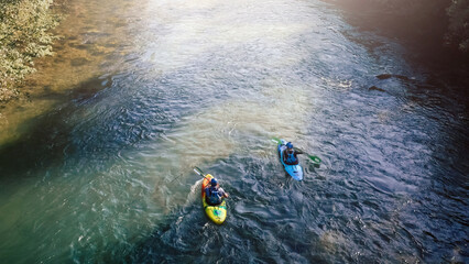 Two male kayakers paddling one after the other, descending a mountain river, aerial rear shot.
