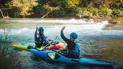 Two smiled Caucasian recreational sportsmen floating in kayaks on the river, giving high five to...