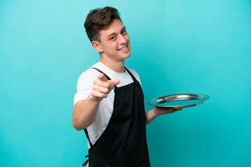 Young waitress with tray isolated on blue background pointing front with happy expression - obrazy, fototapety, plakaty