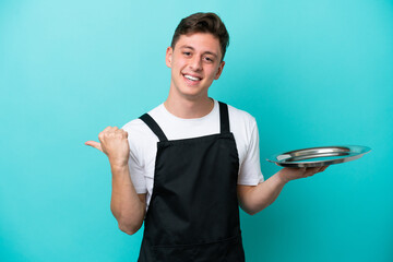 Young waitress with tray isolated on blue background pointing to the side to present a product