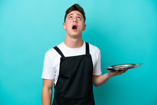 Young waitress with tray isolated on blue background looking up and with surprised expression
