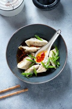 Dark-grey bowl with chili broth and steamed panasian dumplings, vertical shot on a light-blue stone background