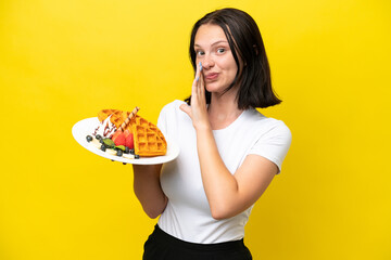 Young caucasian woman holding waffles isolated on yellow background whispering something