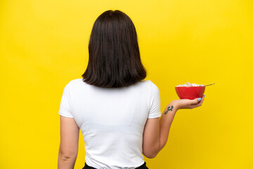 Young caucasian woman holding a bowl of cereals isolated on yellow background in back position