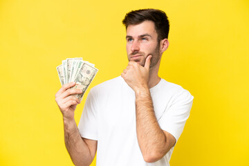 Young caucasian man taking a lot of money isolated on yellow background having doubts