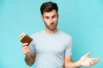 Young caucasian man holding a wallet isolated on blue background making doubts gesture while lifting the shoulders