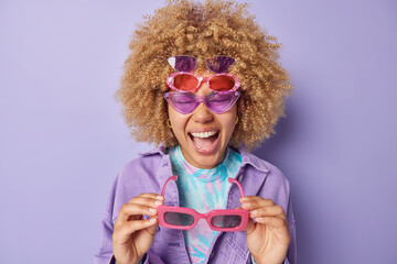 Overjoyed woman tries to choose best sunglasses exclaims loudly prepares for summer season dressed in fashionable jacket isolated over purple background. Hard choice concept. Which one better fit me