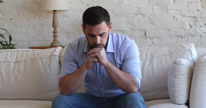 Stressed concerned millennial man thinking over bad news, problem solving, sitting on couch at home, touching, rubbing face, leaning chin on hands, feeling depressed, frustrated, coping with crisis