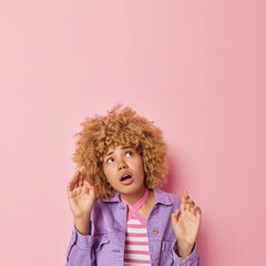 Scred woman keeps hands like something falling from above tries to protect herself from danger focused overhead wears purple jacket isolated over pink background empty space for your advert.