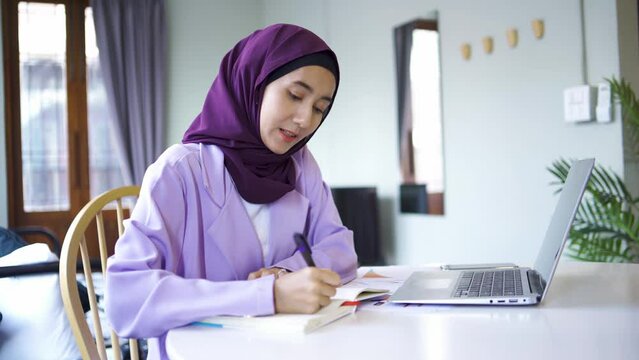A Muslim woman wearing a hijab sits at a table in a cafe working with a laptop and a notepad.