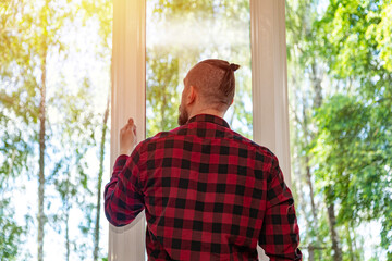 man in red plaid shirt opens white plastic window. nature background. clean air