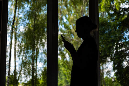 Silhouette of woman holding a mobile phone while standing near glass window