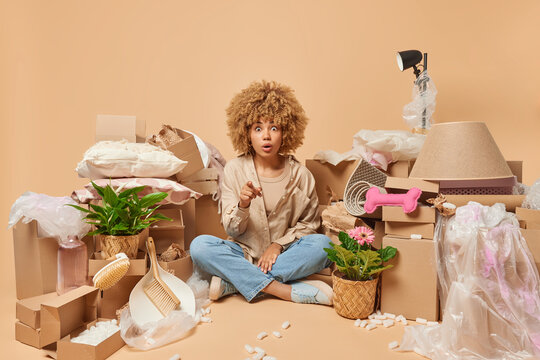 Shocked amazed woman with curly hair sits crossed legs wears shirt and jeans poses around carton boxes relocates to new flat points into camera isolayed over beige background. Real estate agency
