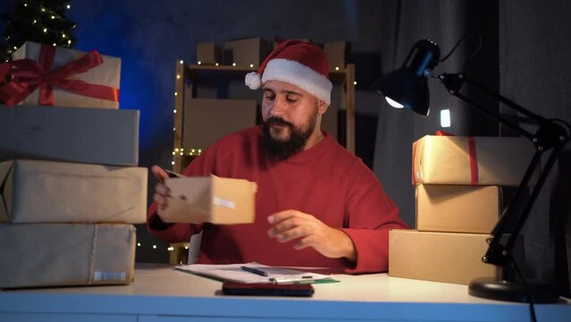 Dropshipping owner Indian man Santa Claus works at night in his home office on laptop, writes the delivery address on the box before sending it to the client during the Christmas sale.