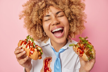 Cheerful female student prefers eating fast food holds delicious hot dog and burger laughs out gladfully wears white formal shirt smeared with ketchup and blue tie isolated over pink background