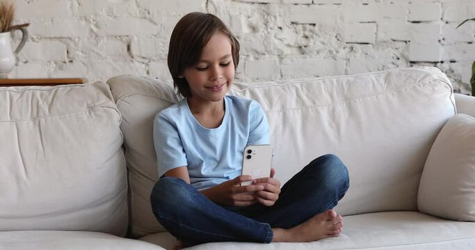 Focused cute preteen child using mobile phone on couch in living room, getting good news, smiling, laughing, looking at screen. Happy gen Z kid sitting on sofa, chatting online, playing videogame