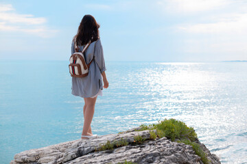 Fototapeta na wymiar a traveler girl in summer clothes with a fashionable backpack on a white rock on the seashore