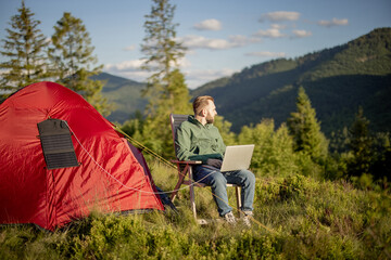 Man works on laptop while traveling with tent in the mountains. Charging computer with portable...