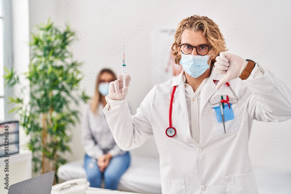 Poster Blond man wearing doctor uniform and medical mask holding syringe with angry face, negative sign showing dislike with thumbs down, rejection concept