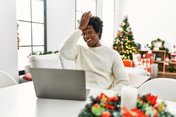 Young african american man using laptop sitting on the table by christmas tree surprised with hand on head for mistake, remember error. forgot, bad memory concept.