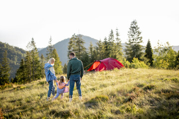 Fototapeta na wymiar Young caucasian family with little girl walk together on green meadow while traveling with tent in the mountains during sunset. Happy family spending summer vacation at campsite