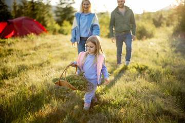 Little girl walks with basket full of wildflowers her parents running behind. Young family happily spending summer time traveling in the mountains