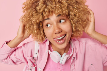 Dreamy teenage girl sticks out tongue looks away pensively keeps hands on her curly bushy hair wears fashionable jacket and headphones around neck for listening music isolated over pink background.