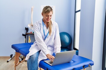 Young blonde woman wearing physiotherpist uniform using laptop at physiotherapy clinic