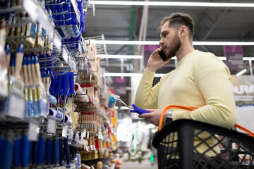 a man in a hardware store chooses paint brushes while discussing it on the phone with his wife