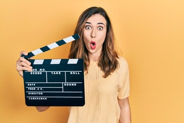 Young hispanic girl holding video film clapboard scared and amazed with open mouth for surprise,...