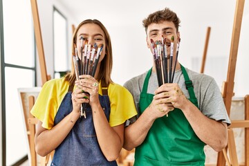 Young hispanic artist couple smiling happy covering mouth with paintbrushes at art studio.