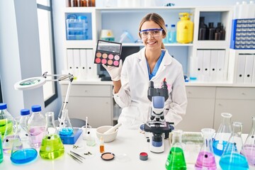 Young blonde woman working at scientist laboratory with make up looking positive and happy standing and smiling with a confident smile showing teeth