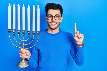 Young hispanic man holding menorah hanukkah jewish candle smiling with an idea or question pointing...