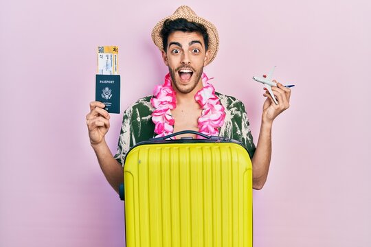 Young hispanic man wearing summer style and hawaiian lei holding passport and plane toy celebrating crazy and amazed for success with open eyes screaming excited.