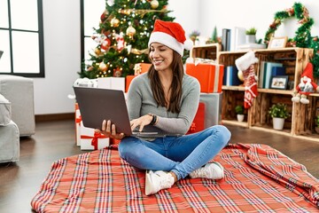 Young latin woman using laptop sitting by christmas tree with a happy and cool smile on face. lucky person.