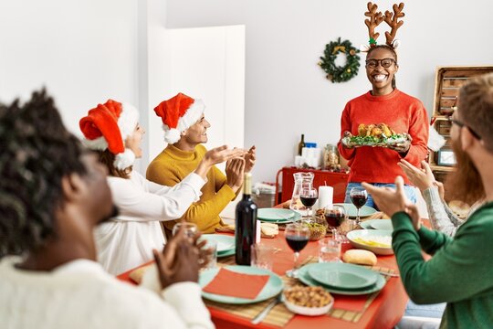 Group of people meeting clapping and sitting on the table. Woman standing and holding roasted turkey celebrating Christmas at home.