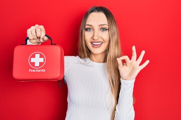 Young blonde girl holding first aid kit doing ok sign with fingers, smiling friendly gesturing...